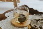 Glastasse mit Teesieb auf einem Holztablett neben einer Glaskanne mit Kräutertee