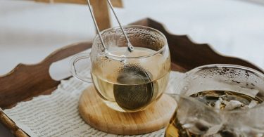 Glastasse mit Teesieb auf einem Holztablett neben einer Glaskanne mit Kräutertee