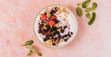 Schale mit Joghurt, Beeren, Nüssen und Minzblättern auf rosa Untergrund, von oben gesehen