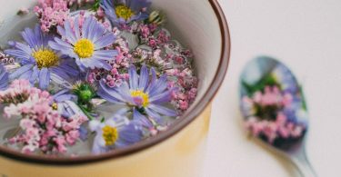 Schale mit Wasser und lila und rosa Blüten, daneben ein Löffel mit Blüten