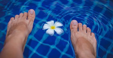 Füße im klaren Wasser eines Schwimmbeckens, dazwischen treibt eine weiße Blüte