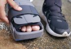 Fuß in einer Orthese neben einem Sportschuh, eine Hand schließt den Klettverschluss