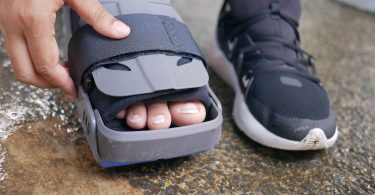 Fuß in einer Orthese neben einem Sportschuh, eine Hand schließt den Klettverschluss