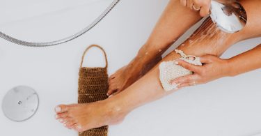 Wasserstrahl aus einer Handbrause über ein Bein, daneben Badebürste und Waschlappen