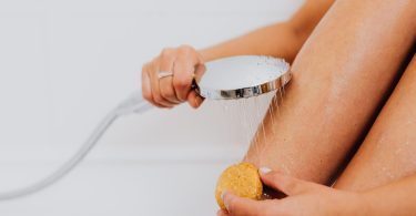 Hand führt eine Handbrause über ein Bein, die andere hält einen Naturschwamm