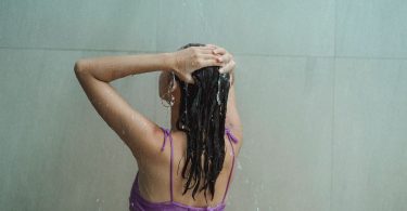Frau steht von hinten gesehen unter der Dusche und hält die Hände am nassen Haar