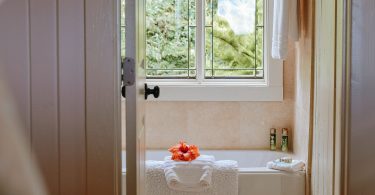 Blick durch eine offene Tür auf eine Badewanne mit Handtüchern und einer Blüte vor einem Fenster