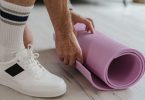 Person in Turnschuhen rollt eine rosa Gymnastikmatte auf einem Holzboden