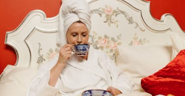 Frau im Bademantel mit Handtuch um den Kopf trinkt im Bett eine Tasse Tee