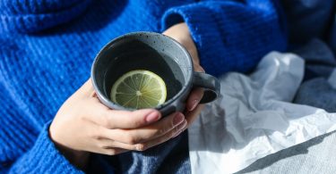 Hände halten einen Becher mit heißem Zitronenwasser, daneben Taschentücher