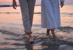 Zwei Menschen waten barfuß mit hochgekrempelten Hosen durch flaches Wasser am Strand