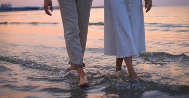 Zwei Menschen waten barfuß mit hochgekrempelten Hosen durch flaches Wasser am Strand